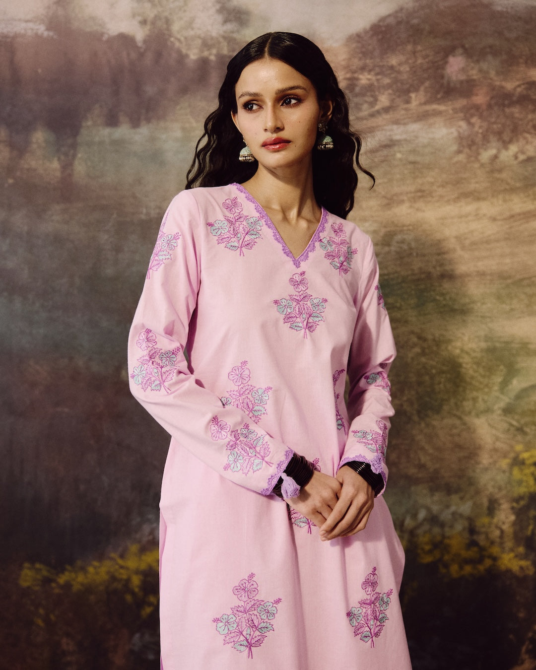 A woman modeling a light pink long-sleeve kurta with purple and green floral embroidery, set against a rustic, textured brown and beige background.